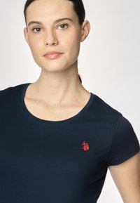 Una donna con la pelle liscia e i capelli scuri indossa una t-shirt aderente blu navy decorata con un piccolo logo rosso, sorridendo dolcemente su uno sfondo chiaro.