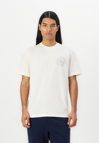 Tricou alb din bumbac, cu guler rotund și mâneci scurte, având un logo circular gri pe partea stângă a pieptului. Textură netedă, croială lejeră.
