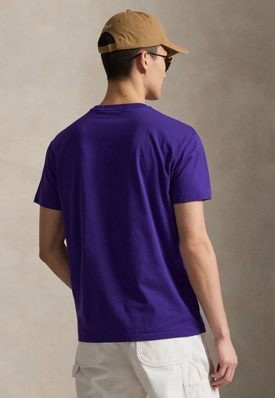 T-shirt en coton violet avec un col rond, des manches courtes et une texture lisse, porté avec un pantalon de couleur claire et une casquette beige.