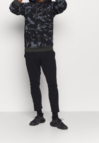 Czarna hoodie w kamuflażu z ściągaczem, zestawiona z czarnymi spodniami slim-fit i czarnymi butami sportowymi. Miękki materiał, luźny krój i ściągacze na mankietach.