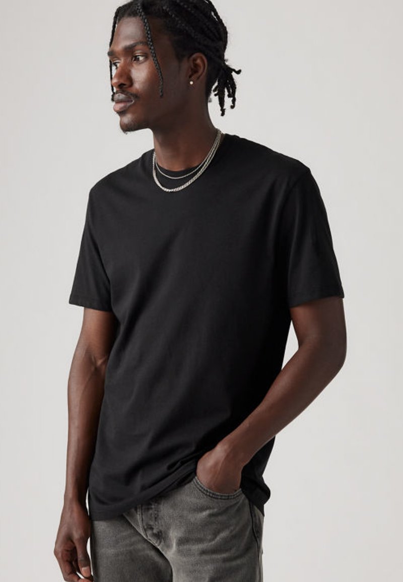 Juoda medvilninė marškinėliai su apvalia iškirpte, trumpomis rankovėmis ir tiesia apačia. Modelis dėvi pilkus džinsus ir sluoksniuotas sidabrines apyrankes.