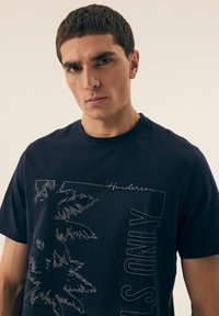 Granatowa bawełniana koszulka z grafiką palm, wzorem w kropki i czarnymi napisami "Henderson" oraz "IS ONLY." Okrągły dekolt, krótkie rękawy.