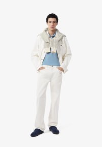 Biała dżinsowa kurtka, jasnoniebieski sweter, kremowy dziany szalik, białe spodnie, ciemnoniebieskie sneakersy. Model pozuje z rękami w kieszeniach.