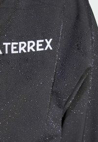 Veste noire résistante à l'eau arborant le logo "WATERREX" en blanc, avec des gouttes d'eau visibles sur la surface texturée du tissu.