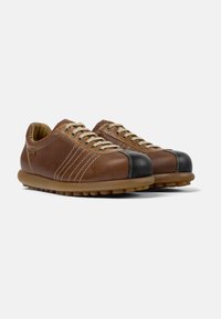 Scarpe in pelle marrone con suola in gomma, dotate di una punta nera arrotondata, cuciture a contrasto e lacci beige. Dettaglio del materiale testurizzato.