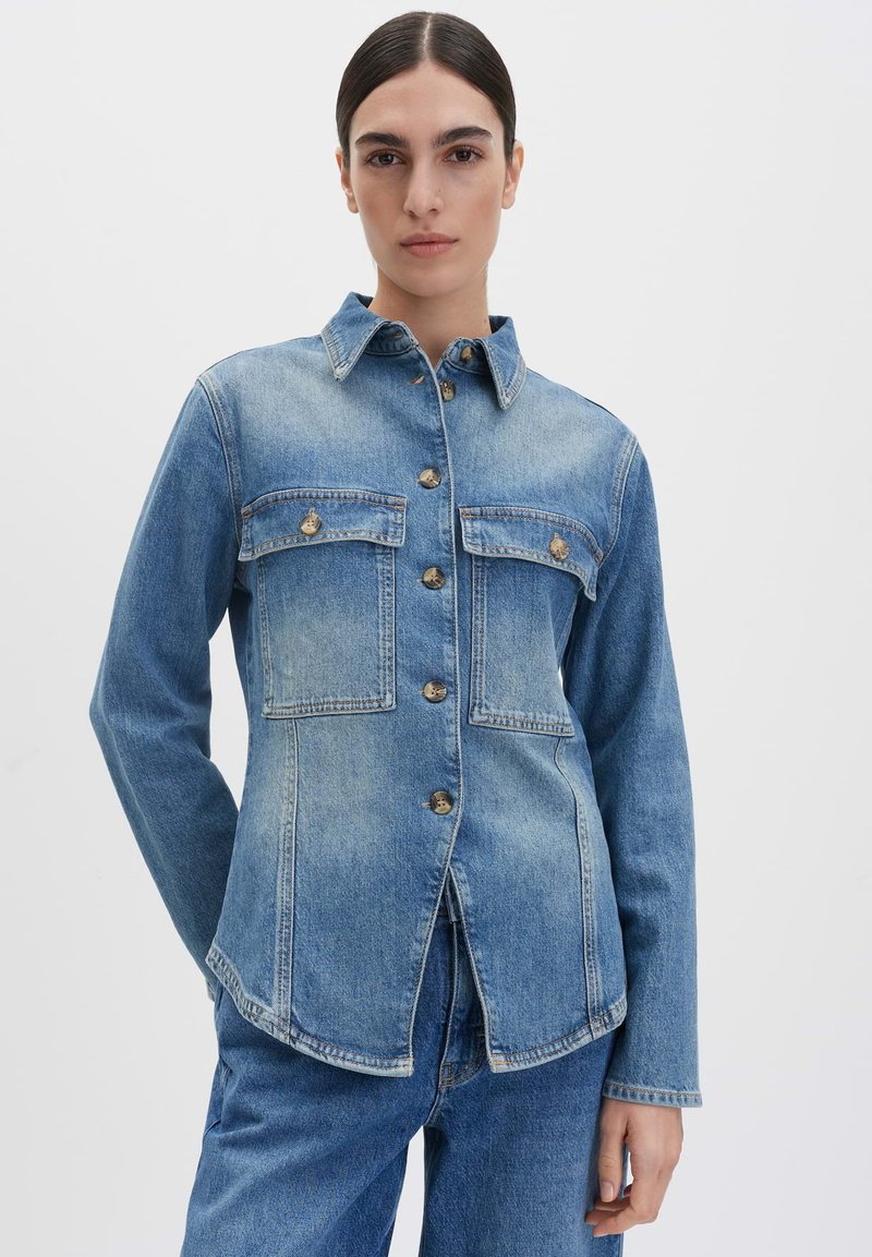 Giacca di jeans blu con chiusura a bottoni, colletto a punta, due tasche sul petto e maniche lunghe. Presenta un orlo curvo e un taglio classico.