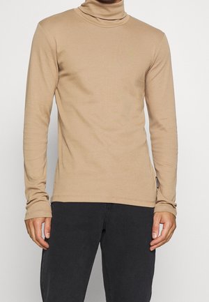 Homme portant un col roulé à manches longues beige et un pantalon noir, debout devant un fond clair uni.