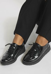 Chaussures brogue noires en cuir verni avec semelles épaisses, portées avec un pantalon noir court sur fond clair.