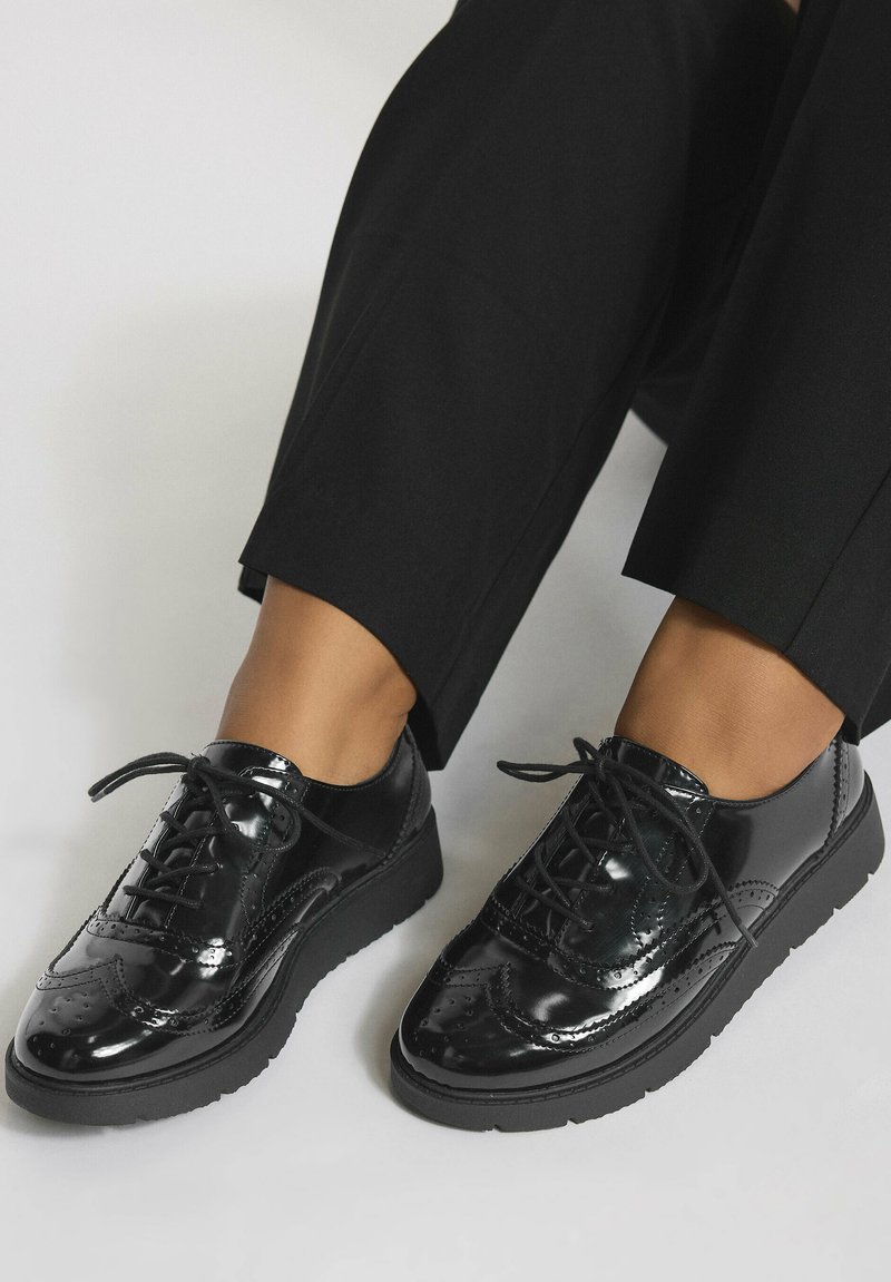 Chaussures brogue noires en cuir verni avec semelles épaisses, portées avec un pantalon noir court sur fond clair.