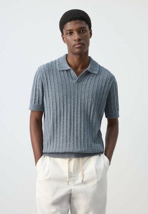 Joven con cabello corto y rizado que lleva un polo azul de manga corta de punto acanalado y pantalones blancos con cordón, con las manos en los bolsillos.