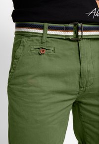 Chinos en coton vert avec un tissu texturé, ornés d'un bouton marron et d'une ceinture tissée à motifs dans des couleurs vert, marine, blanche et marron.