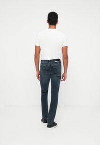 Vyras stovi atsukęs nugarą, vilkėdamas baltą marškinėlį, tamsiai mėlynas džinsus ir juodus sportbačius prieš vienodą baltą foną.