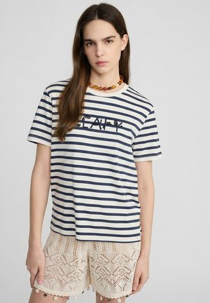 Joven mujer con camiseta de rayas azul marino y blanca con el texto "HAPPY" y pantalones cortos de encaje de ganchillo beige, de pie frente a un fondo liso.