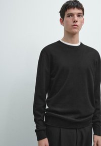 Schwarzer gestrickter Pullover mit Rundhalsausschnitt, langen Ärmeln, gerippten Bündchen und Saum. Über einem weißen Hemd getragen, mit glatter Textur.