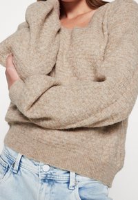 Frau in einem beigen strukturierten Strickpullover mit langen Ärmeln, der über der Brust gekreuzt ist, kombiniert mit hellblauen Denim-Jeans.