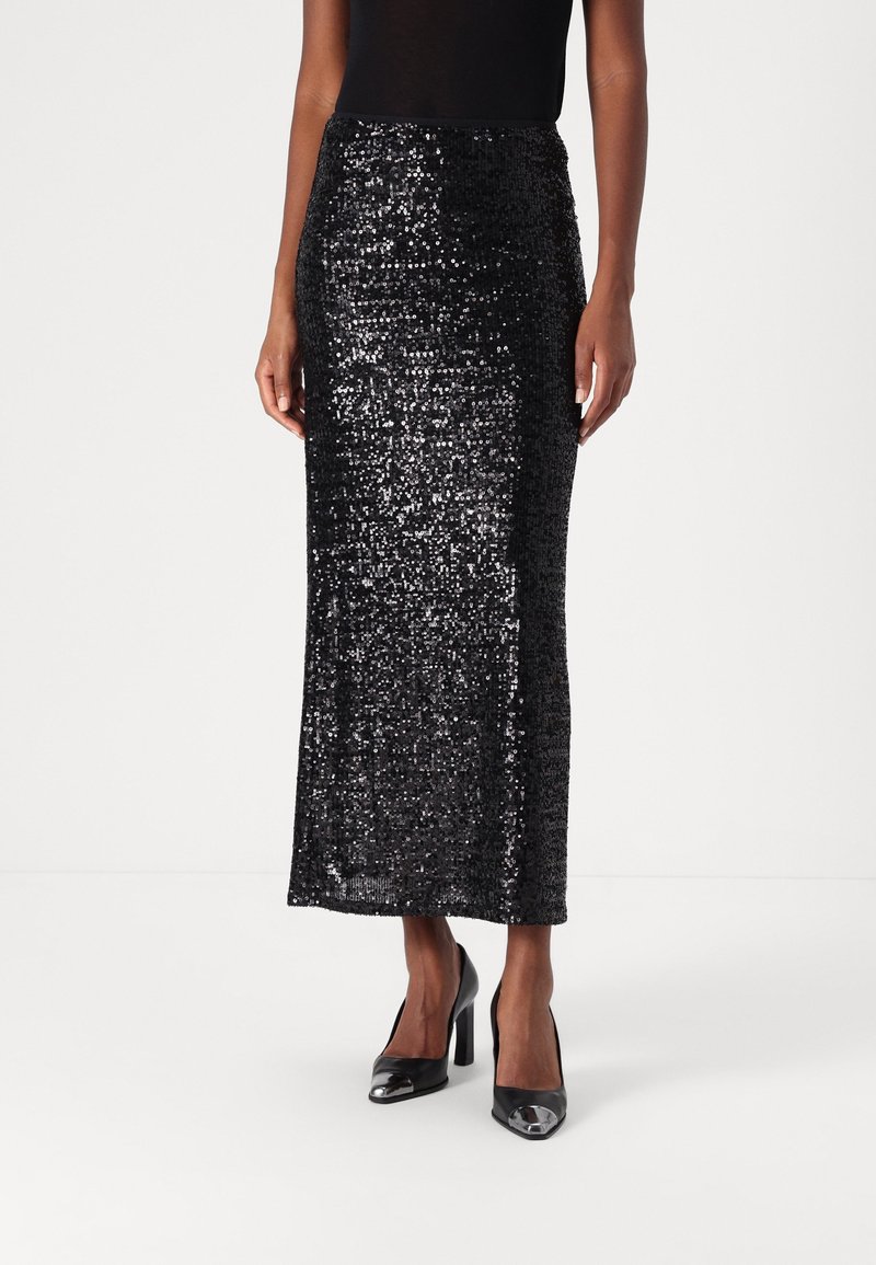 Femme portant une jupe midi noire à sequins associée à des chaussures à talons hauts noires brillantes, debout devant un fond blanc uni.