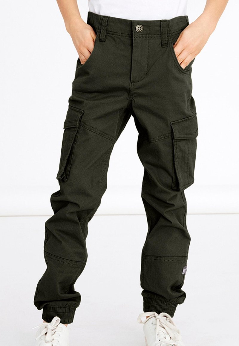 Enfant portant un pantalon cargo vert foncé avec des poignets élastiques et des baskets blanches, mains dans les poches avant, debout sur un fond blanc.