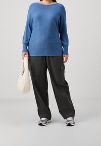 Maglione blu a coste, vestibilità rilassata, maniche lunghe. Pantaloni cargo neri con tasche laterali. Una borsa bianca e scarpe da ginnastica grigie completano il look.