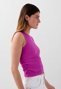 Top aderente senza maniche in materiale a righe di un vivace rosa, con scollatura a girocollo e dettagli arricciati sui lati, abbinato a pantaloni bianchi.
