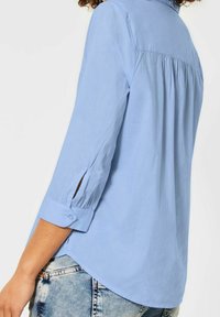 Světle modrá košile s dlouhým rukávem, s manžetou na knoflík, zadními sklady a bočními rozparky. Vyrobena z lehkého materiálu. Spodní lem je zakřivený.