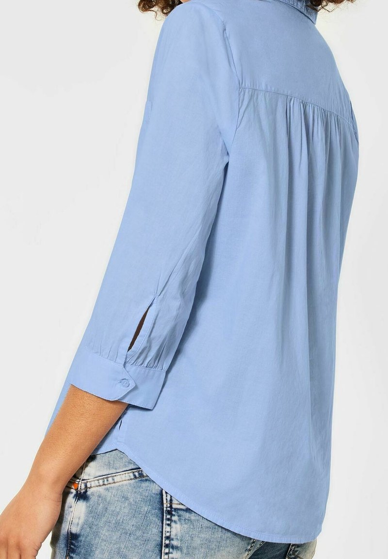 Světle modrá košile s dlouhým rukávem, s manžetou na knoflík, zadními sklady a bočními rozparky. Vyrobena z lehkého materiálu. Spodní lem je zakřivený.