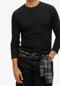 Man som bär en svart långärmad skjorta och mörka jeans, med en grå rutig skjorta knuten runt midjan och en svart klocka på vänster handled.