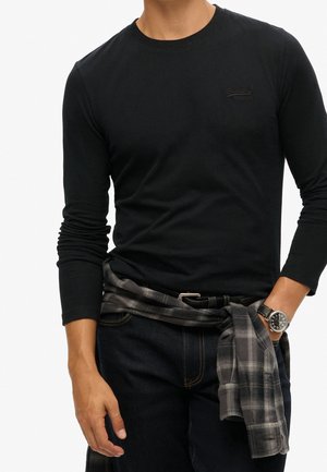 Man in een zwart shirt met lange mouwen en donkere jeans, met een grijs geruit shirt om de taille geknoopt en een zwarte horloge om de linkerpols.