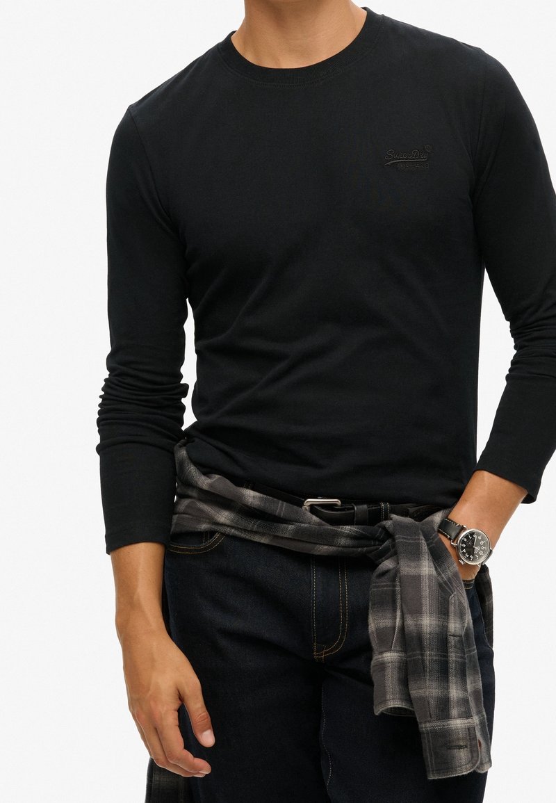 Man som bär en svart långärmad skjorta och mörka jeans, med en grå rutig skjorta knuten runt midjan och en svart klocka på vänster handled.