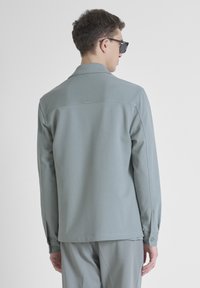 Camicia a maniche lunghe verde chiaro con colletto classico, tessuto testurizzato e polsini con bottoni. Presenta un piccolo logo sulla parte superiore della schiena.