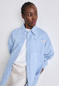 Lys blå denimskjorte med knappestolpe foran, oversized passform, og en enkelt brystlomme med et Calvin Klein-merke.