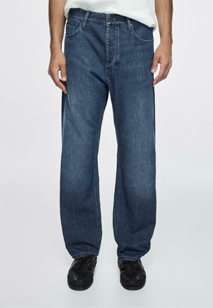 Mann trägt locker sitzende, dunkelblaue Jeans und schwarze Schnürschuhe, steht vor einem einfarbigen hellen Hintergrund.