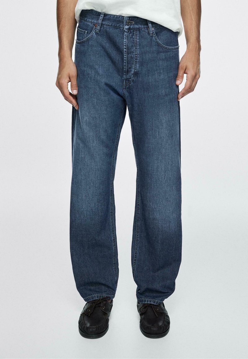 Muž nosící volné tmavě modré džíny a černé šněrovací boty, stojící před čistým světlým pozadím.