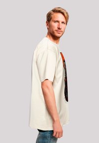 Cremfarbener T-Shirt mit kurzen Ärmeln, das ein farbenfrohes Grafikdesign auf der Vorderseite zeigt. Das Modell trägt blaue Jeans und steht seitlich.