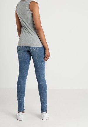 Kobieta w szarym topie bez rękawów, niebieskich obcisłych dżinsach z małymi wzorami i białych trampkach Reebok stoi na tle gładkiej ściany.