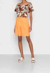 Top floreale senza spalline con fiori rossi e arancioni, abbinato a pantaloni corti plissettati arancioni e sandali beige su uno sfondo neutro.