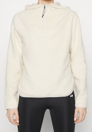 Cremefarvet fleece-pullover med halvlængde lynlås og hætte, båret sammen med sorte leggings med lille hvidt logo nær hoften.