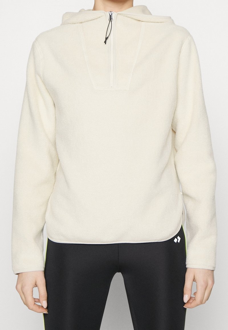 Pull en polaire de couleur crème avec demi-fermeture éclair et capuche, porté avec des leggings noirs présentant un petit logo blanc près de la hanche.