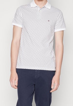 Polo en coton blanc avec motif circulaire noir, patte de boutonnage à trois boutons et col blanc. Porté avec un pantalon bleu marine.