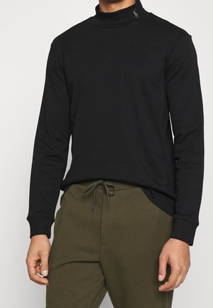 Homme portant une chemise noire à manches longues à col montant factice et un pantalon vert olive à cordon, debout devant un fond clair uni.