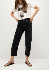 Czarne dżinsy wysokiego stanu o prostym kroju i postrzępionych nogawkach, w zestawieniu z białą koszulką z czarnymi nadrukami graficznymi oraz płaskimi sandałami na paski.