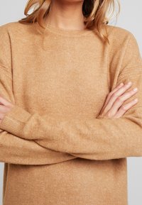 Pull beige en maille avec col rond, manches texturées et poignets côtelés, mettant en valeur un tissu doux et chaud.