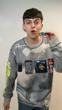 Jeune personne aux cheveux courts et foncés tenant un petit microphone noir avec des lumières vertes près de la bouche, portant un sweat-shirt graphique gris.