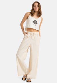 Cropped white tank top with black floral design, paired with loose beige wide-leg pants. Material appears lightweight and breathable.