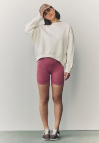 Sudadera blanca extragrande, pantalones cortos rosa con textura acanalada y gorra marrón. La modelo lleva zapatillas marrones y blancas. Diseño de conjunto simple y contemporáneo.