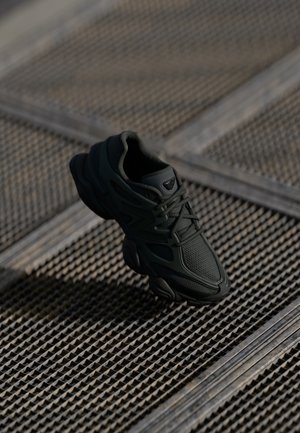 Black athletic sneaker with mesh and synthetic overlays on a textured metal grid surface, lit with soft natural light.