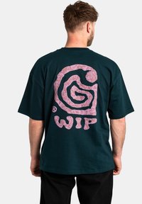 Camiseta de manga corta verde oscuro con un gran logotipo espiral abstracto en rosa y el texto "WIP" en la parte posterior; material de algodón con un corte relajado.