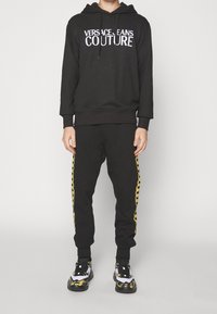 Černá mikina s kapucí s bílým logem "VERSACE JEANS COUTURE", v kombinaci s černými tepláky s žlutými bočními pruhy. Černé a vzorované tenisky.
