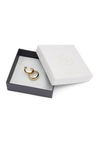 Boucles d'oreilles en or en forme de créole, avec un design lisse et courbé, présentées dans une boîte noire avec un intérieur en velours gris et un couvercle texturé blanc.