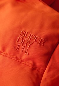 Pomarańczowa kurtka z tkaniny z haftowanym logo "SUPER DRY JPN." Gładka tekstura z widocznymi detalami ściegów na szwie.