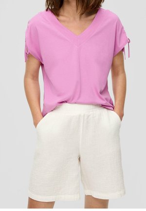 Person iført en løs pink V-hals kortærmet top med bundne ærmedetaljer og hvide teksturerede knælange shorts med lommer.